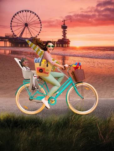 Een vrouw fietst met een kind en strandspullen, op de achtergrond de zee en een reuzenrad.
